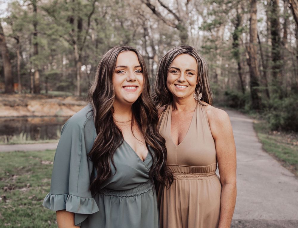 Two people stand close together on a walking path surrounded by trees, wearing a sage green dress and beige dress.
