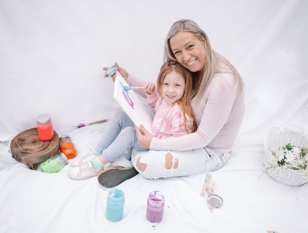 Two people sitting on white floor enjoying playful creative time with art supplies and decorative elements spread around them.