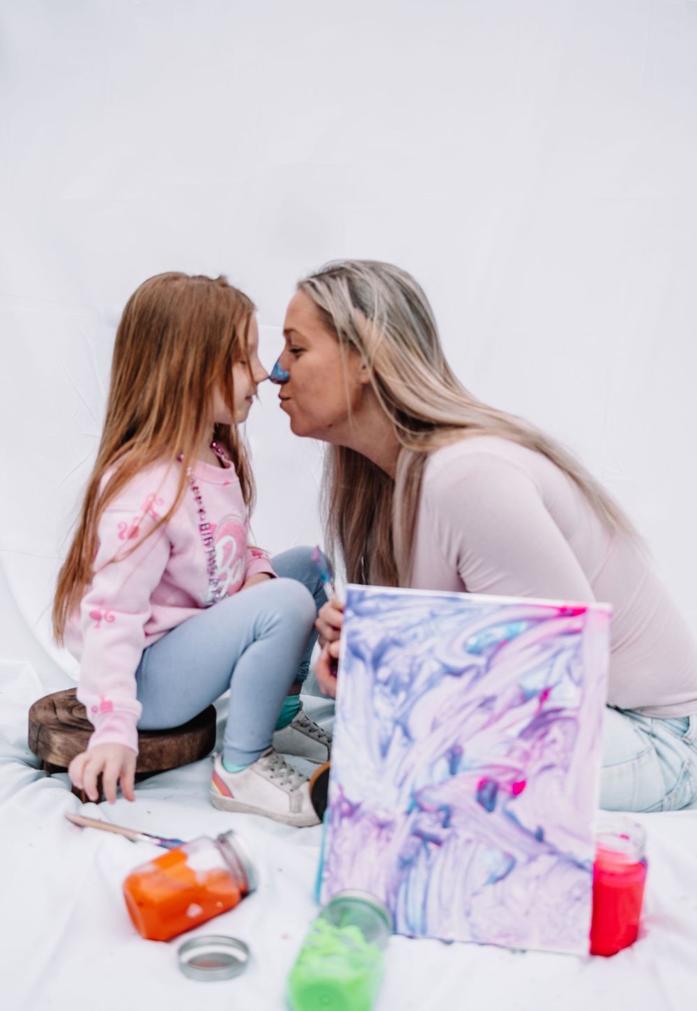 Two people in pink sharing a sweet moment while painting with colorful materials laid out nearby on white surface.