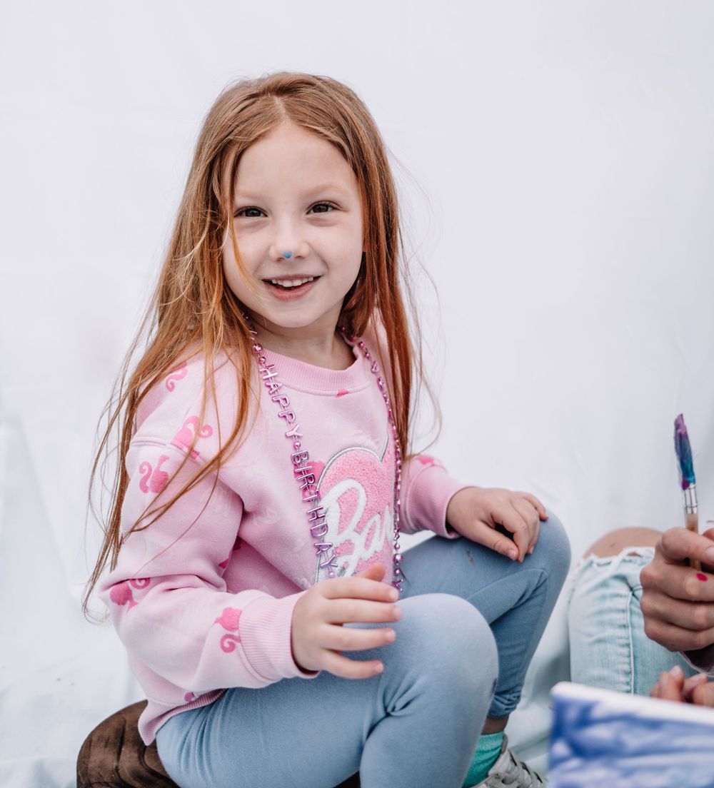 Child in a pink sweater and jeans sits casually while holding something during an outdoor activity.