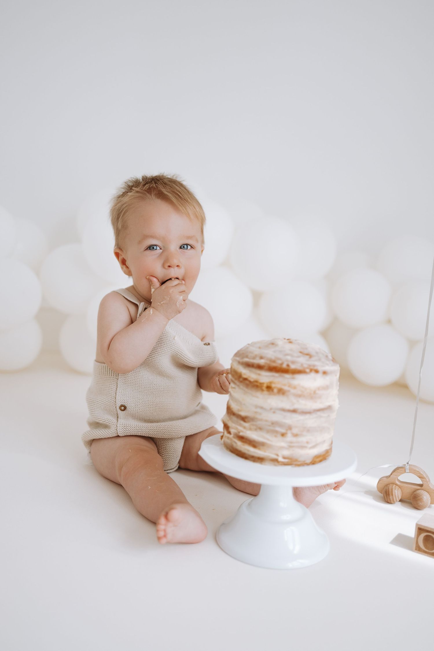 Smiling baby in sage green romper sits beside party balloons and cake during birthday celebration photo session.