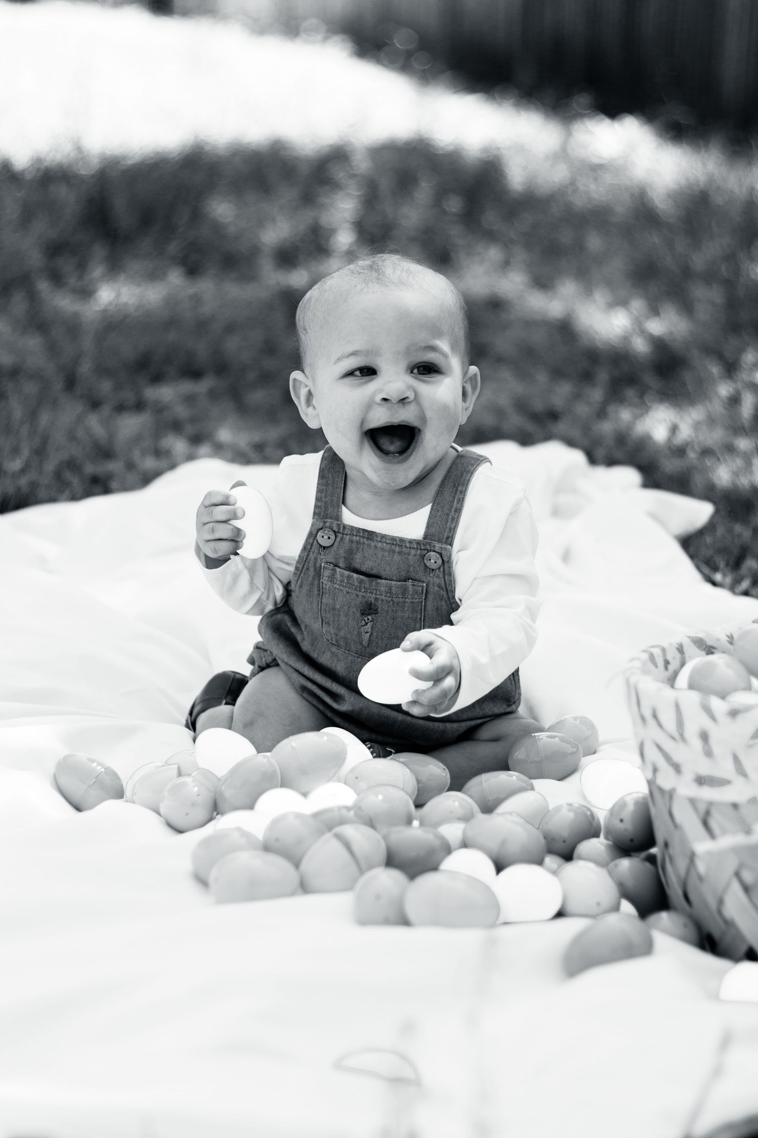 A joyful baby wearing denim overalls sits on a blanket surrounded by fresh oranges in a black and white photograph.