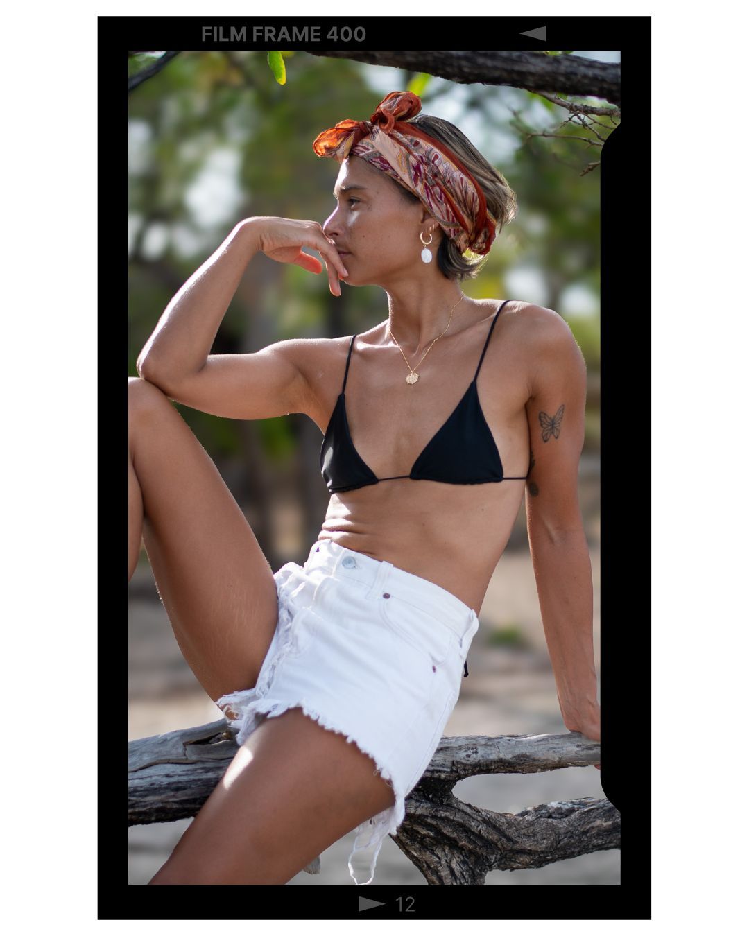 Person in black bikini top and white denim shorts strikes a relaxed pose while sitting outdoors on a log.