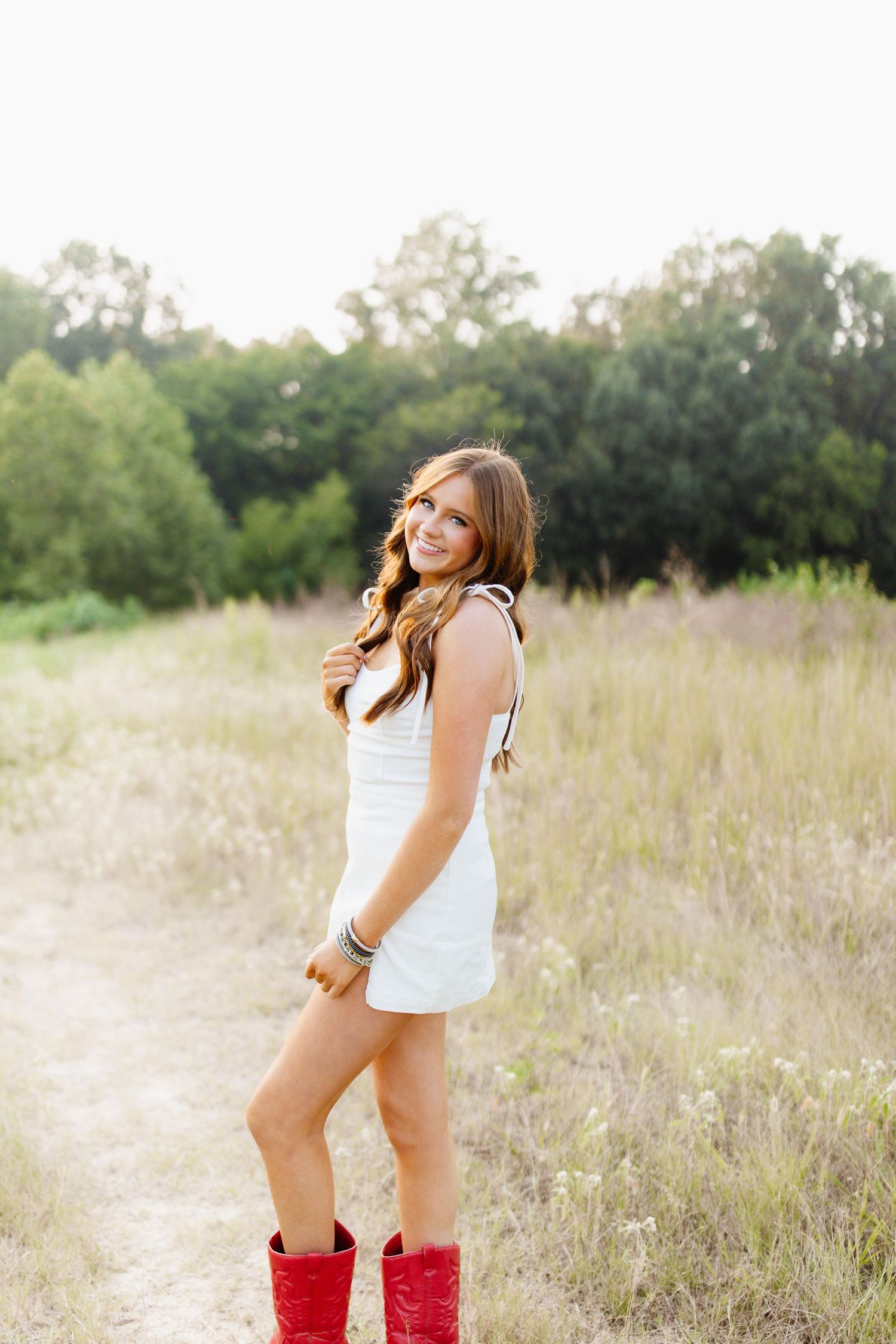A person in a white dress poses in a grassy field while wearing bright red boots.