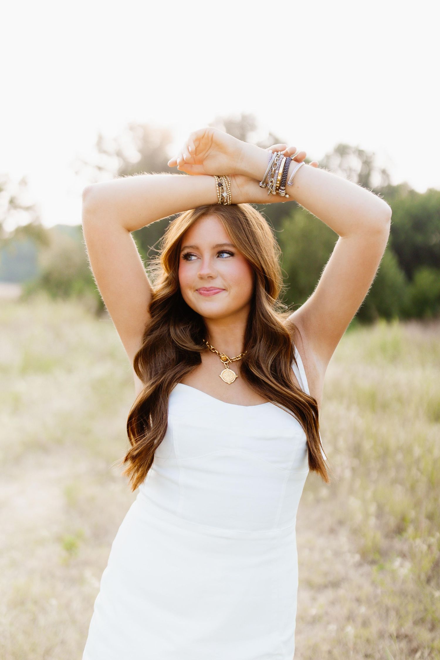 Woman in white dress poses outdoors at golden hour with raised arms and jewelry accessories in a natural field setting.