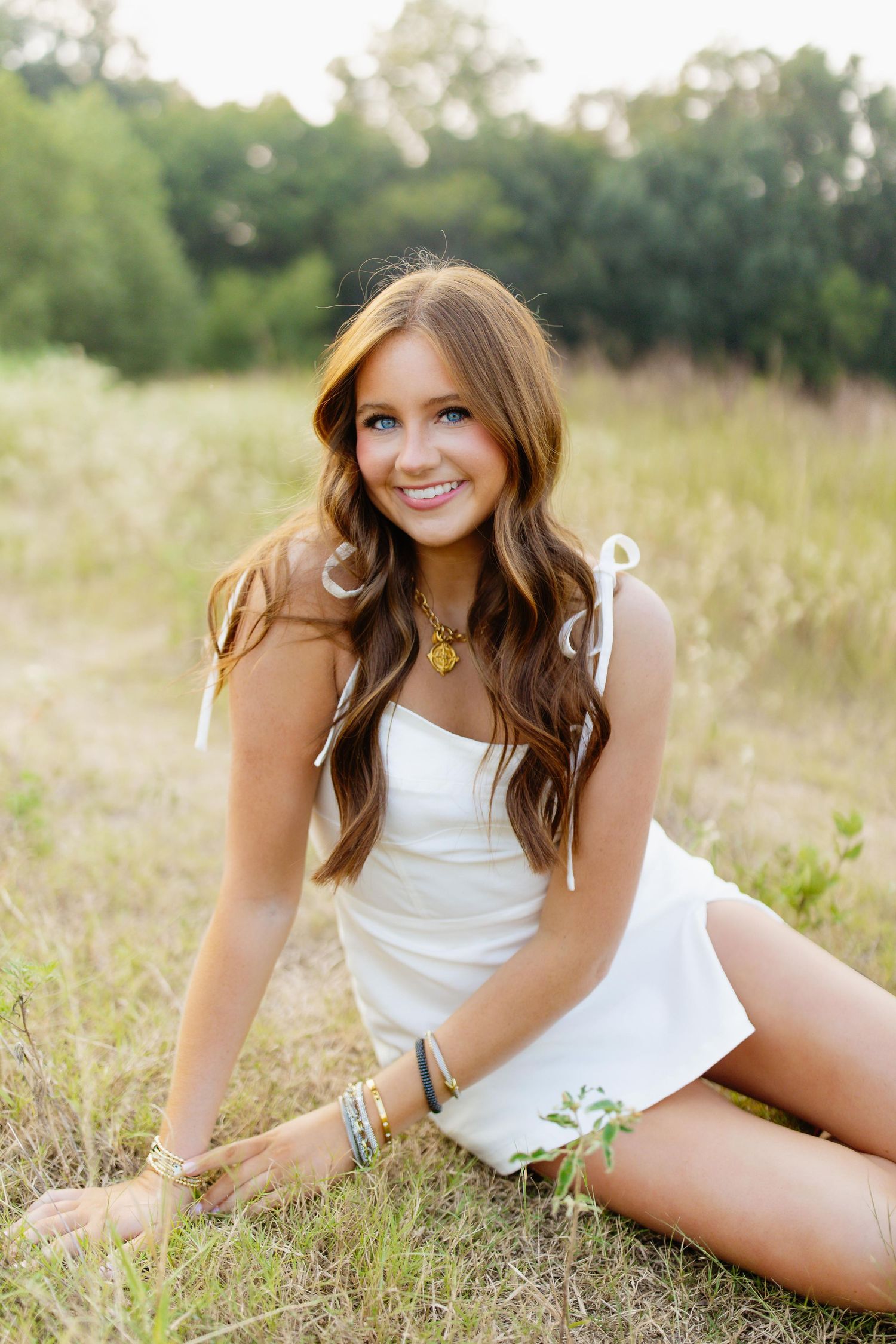 Portrait in a white dress sitting in a golden field at sunset with long wavy brown hair and jewelry.