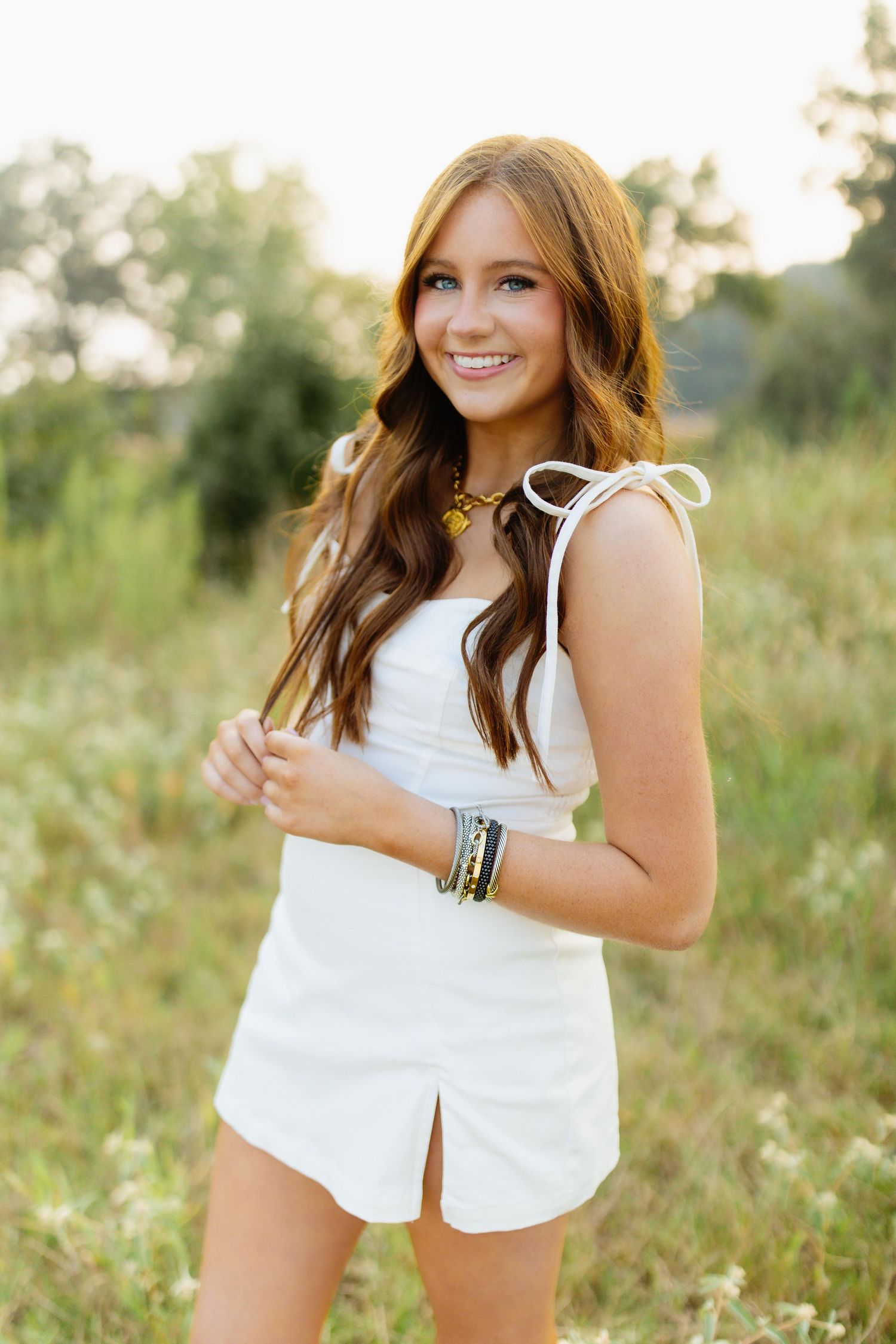 A woman in a white summer romper poses outdoors in a sunny field wearing stacked bracelets.