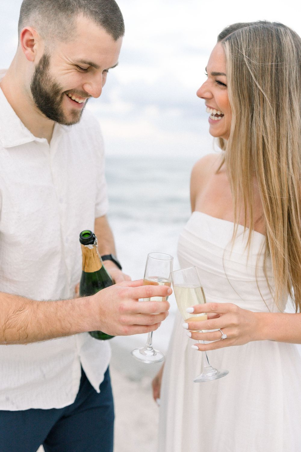 A couple in white outfits shares a romantic moment while toasting with champagne glasses at an outdoor celebration.