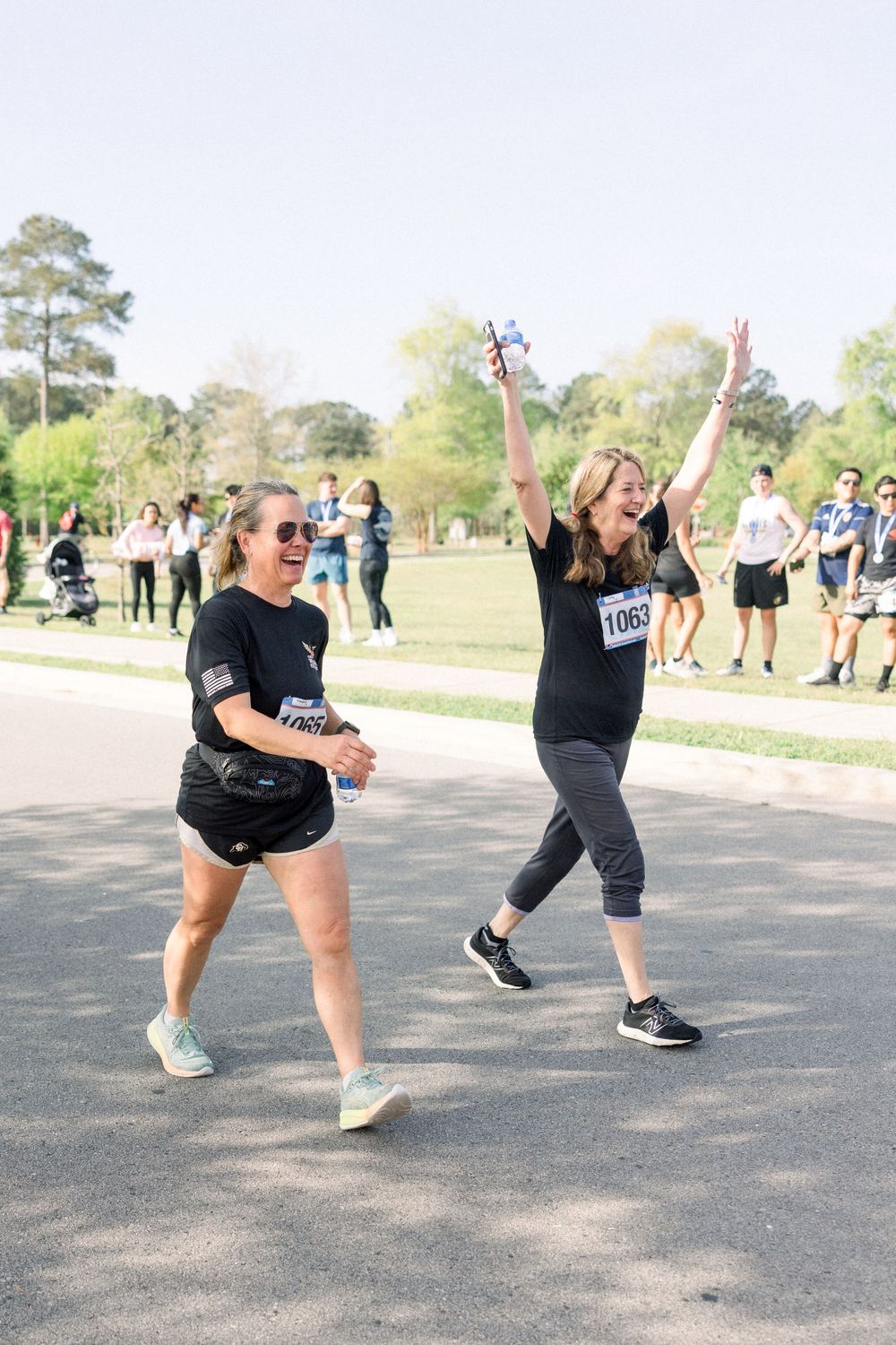 Two participants in athletic wear show excitement while walking during an outdoor running event on a sunny day.
