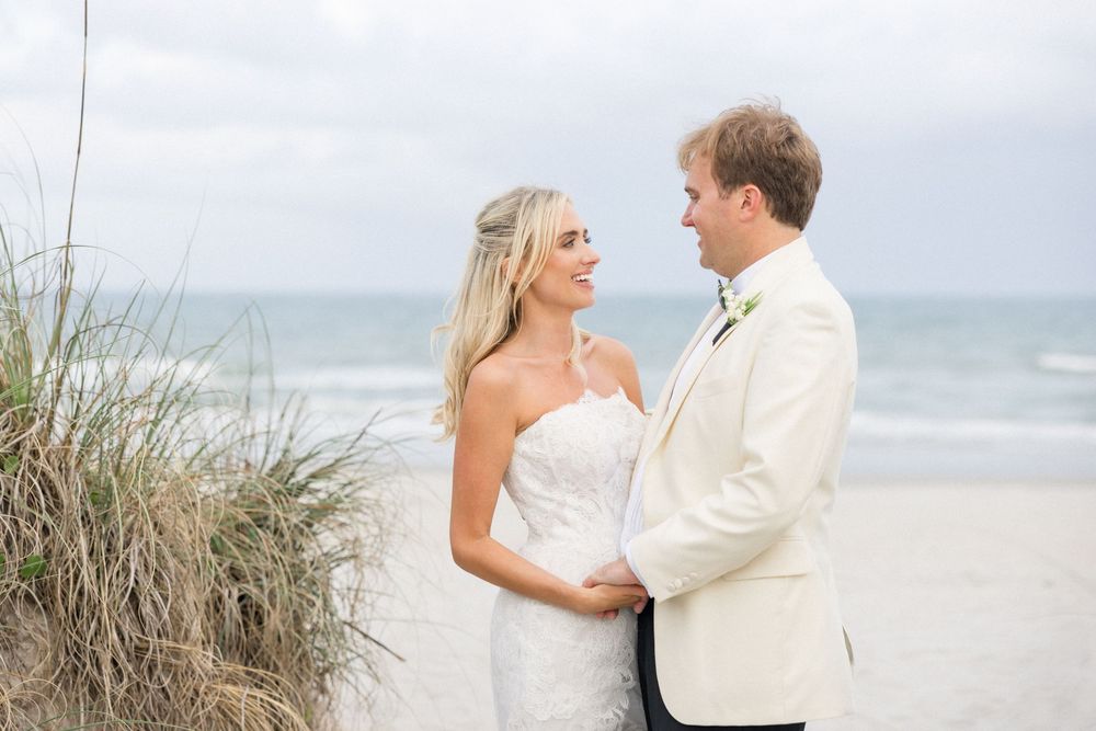 A couple in wedding attire embrace on a sandy beach with sea grass and ocean waves in the background.