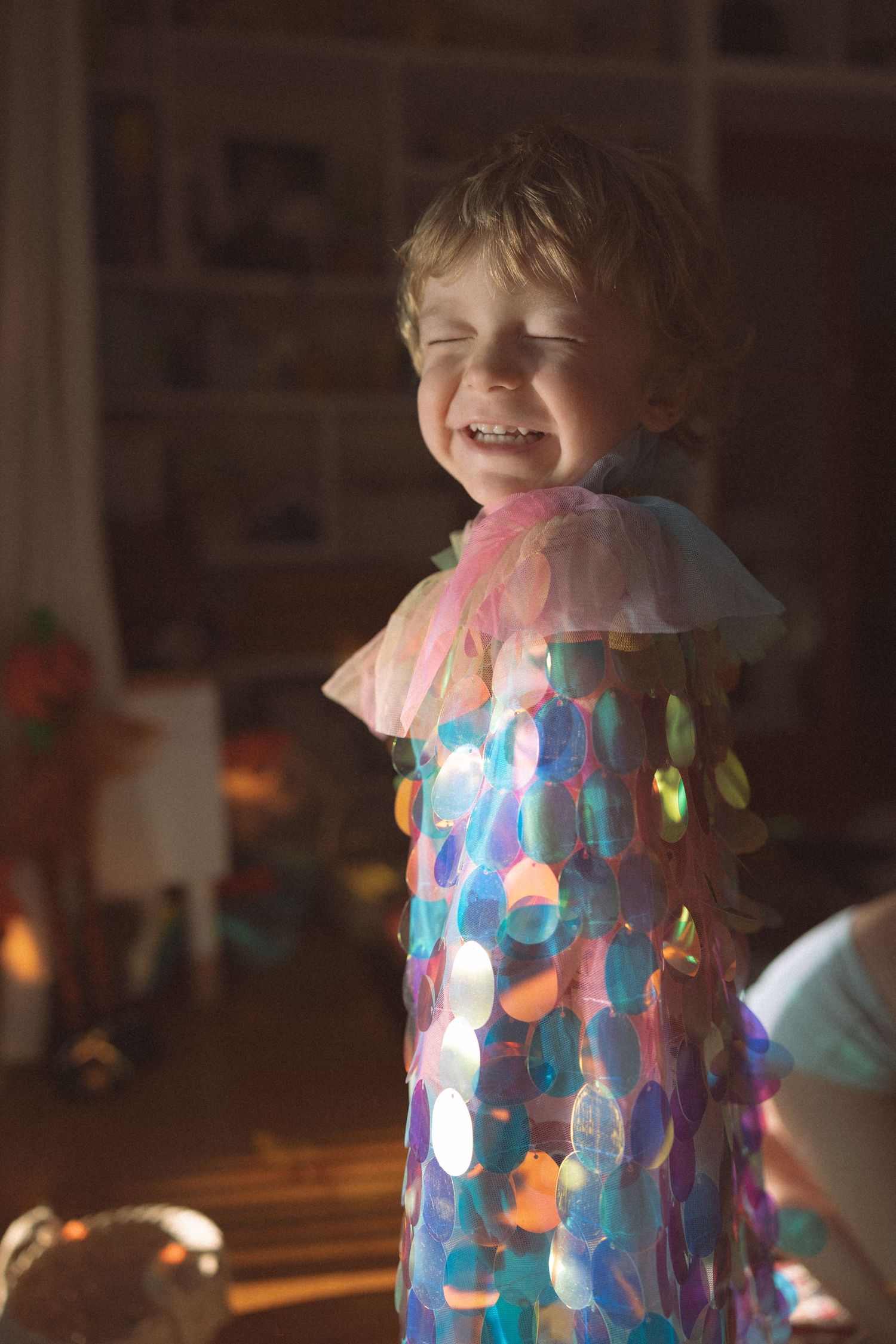 Sunlit room with sheer curtains features flowers scattered on floor while mother and child share tender moment by window.