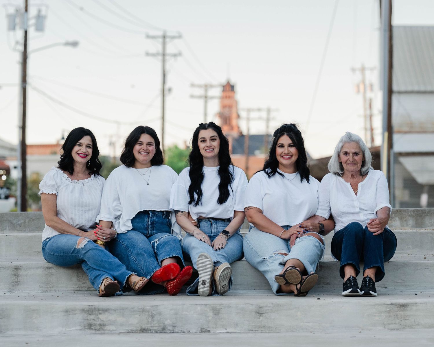 Five women in white tops and jeans sit together on concrete surface at dusk.