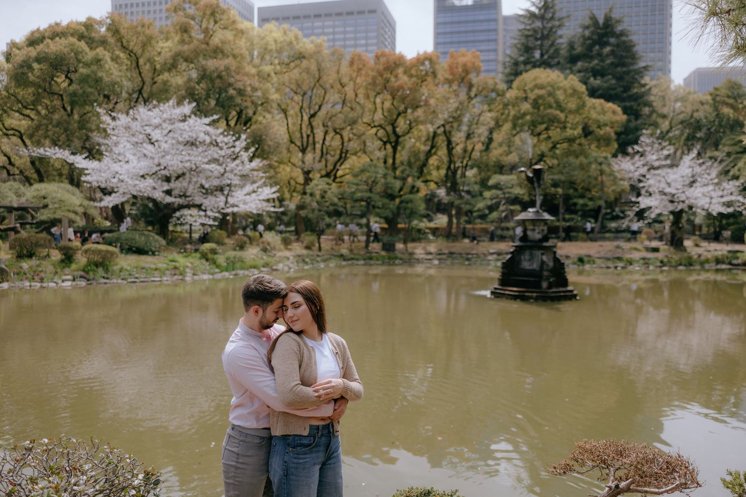 Two people embrace near a peaceful pond surrounded by cherry blossoms and city buildings in a serene park setting.