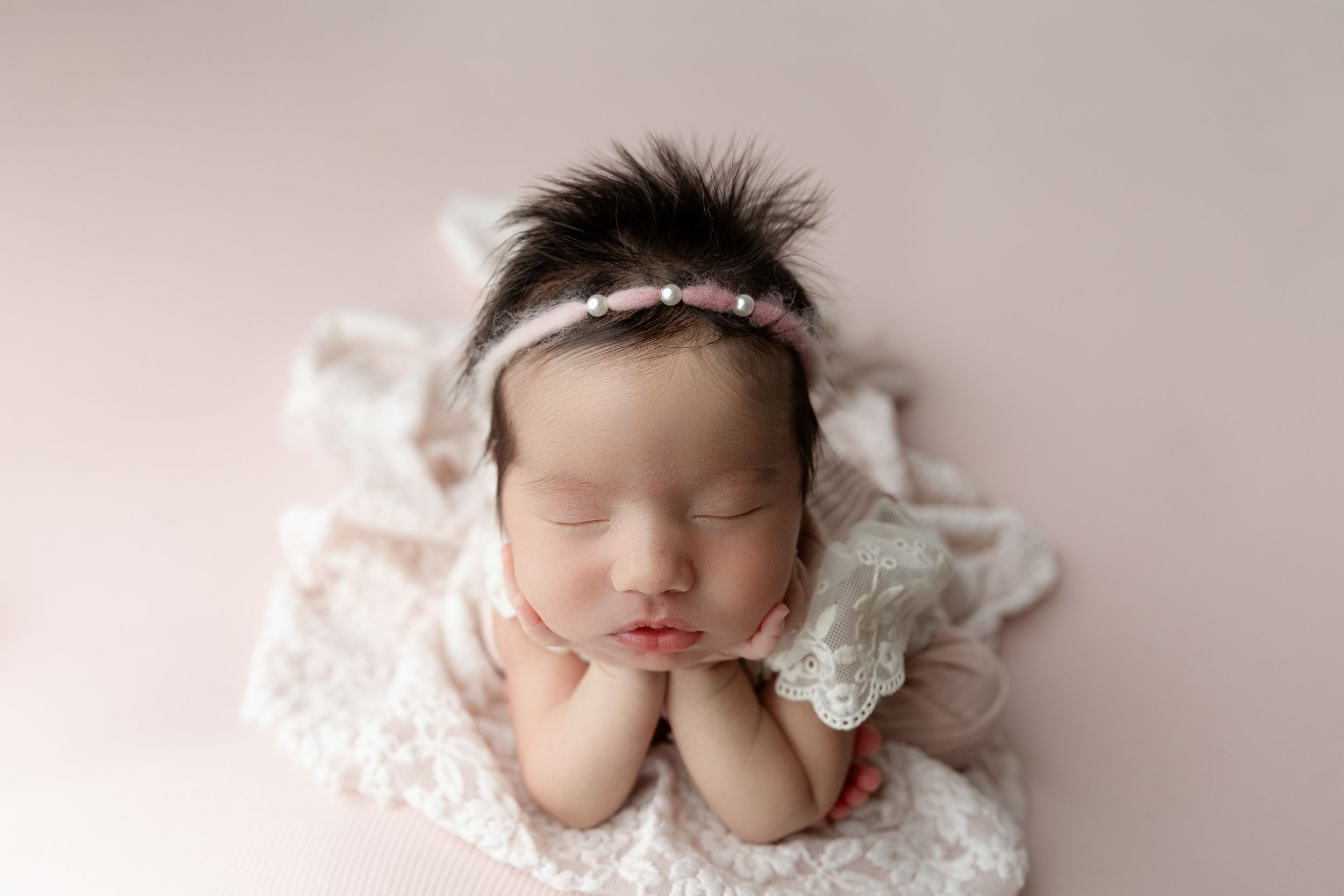 Sleeping newborn on white lace wearing delicate headband in soft pink studio portrait.