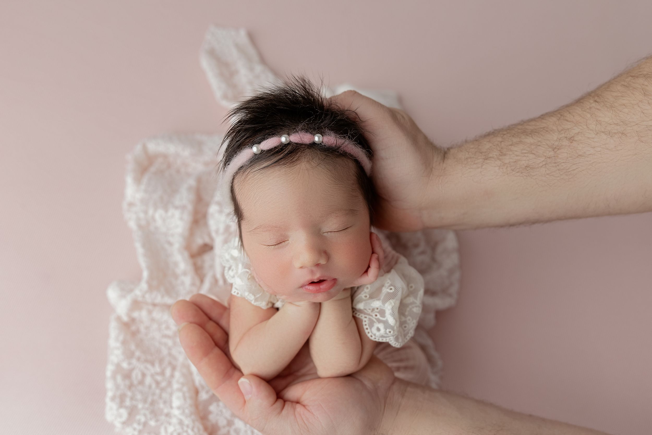 Peaceful sleeping newborn lying on white lace fabric with a delicate headband, supported by gentle hands.