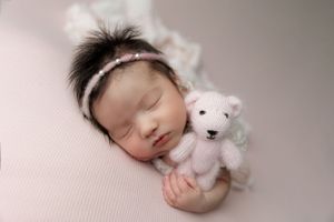 Sleeping newborn in white outfit holding small white teddy bear plush on pink blanket during studio photography session.