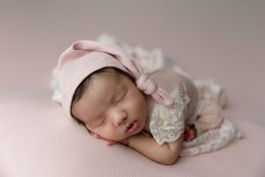 Peaceful sleeping newborn in pink bonnet and white lace dress rests on soft pink backdrop during professional photo session.