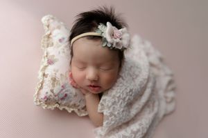 Sleeping newborn wrapped in white lace blanket on pink background with delicate floral headband.