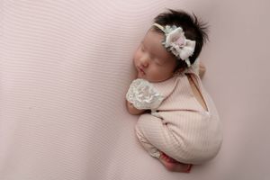 Newborn baby sleeping peacefully in a pink ribbed swaddle with a white floral headband on soft pink backdrop.