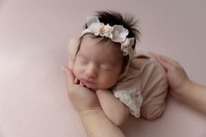 Sleeping newborn on pink blanket wearing white floral headband and lace dress rests peacefully in parent's hands.