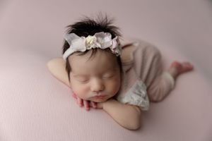 Sleeping newborn in a beige outfit and floral headband rests peacefully on pink fabric backdrop.