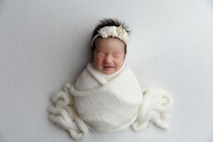 Newborn swaddled in cream white wrap and headband rests peacefully on soft textured blanket during photography session.