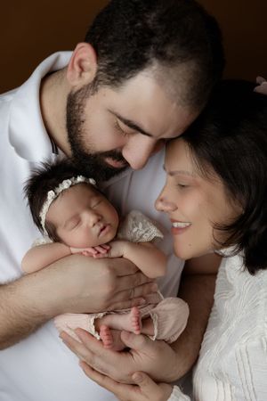 Two parents in white clothing lovingly gaze at their sleeping newborn baby wearing a pearl headband.