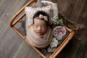 Sleeping newborn wrapped in beige swaddle rests in wooden basket with floral decorations and soft cushion.