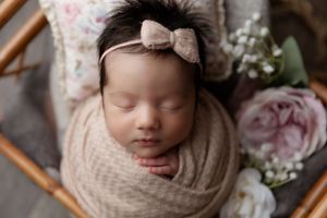 Newborn portrait with a bow headband wrapped in beige fabric next to pink roses and greenery.