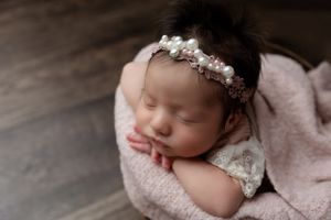 Sleeping newborn wearing delicate lace dress and pearl headband rests peacefully on soft pink blanket.