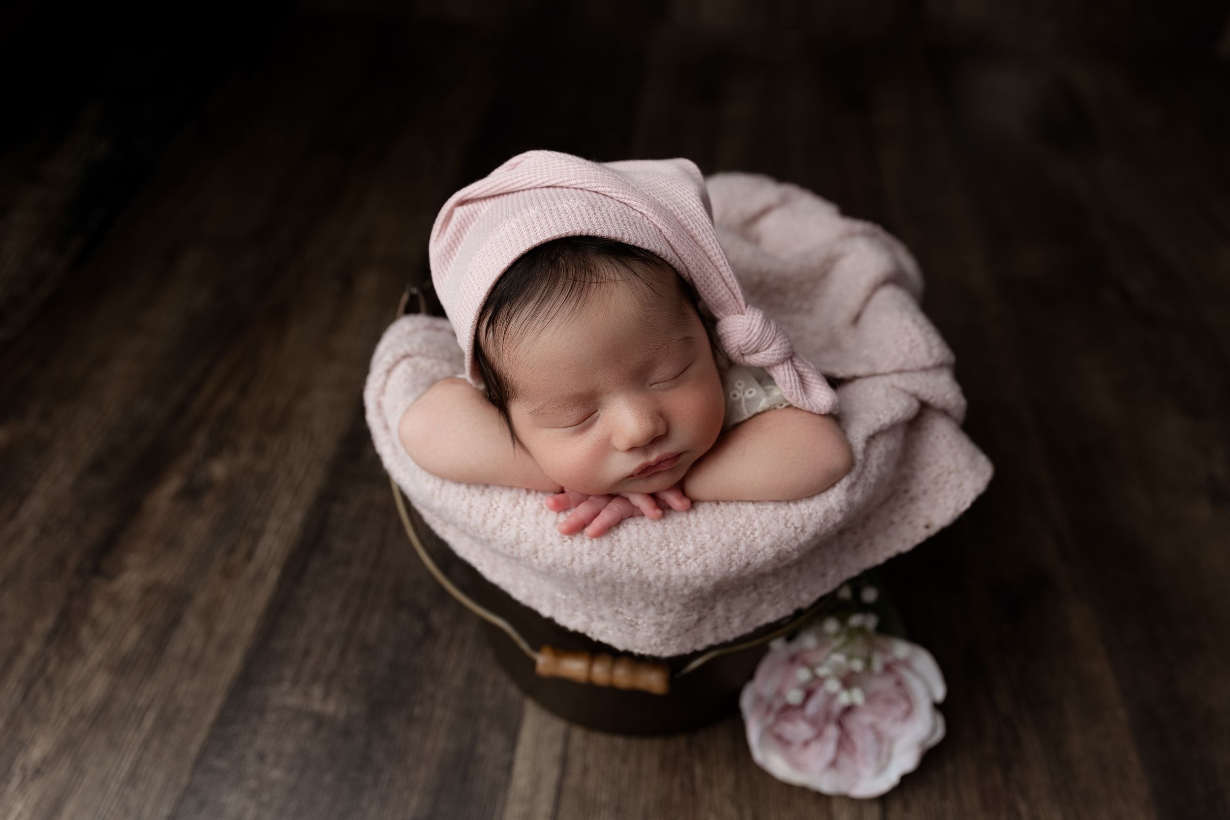 Sleeping newborn in pink swaddle lies peacefully in round basket on wooden floor with small floral decoration.