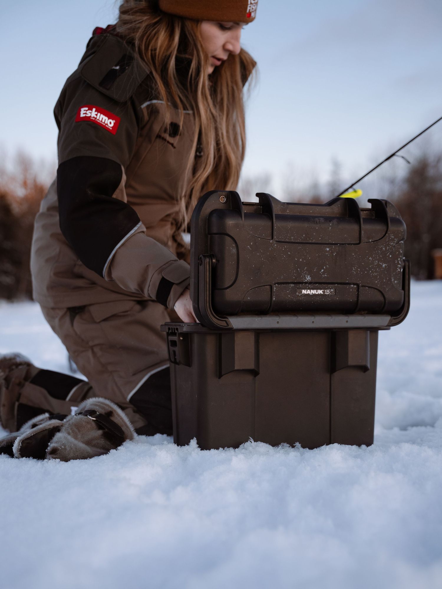 Brown tackle box and fishing equipment arranged in deep snow during ice fishing.
