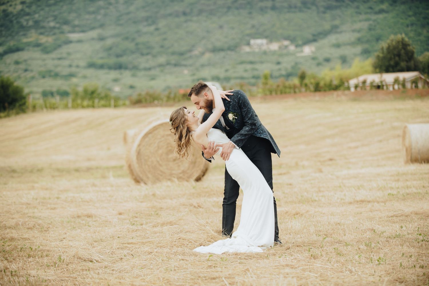 Newlyweds share a romantic dip kiss in a rustic field with hay bales in the background.