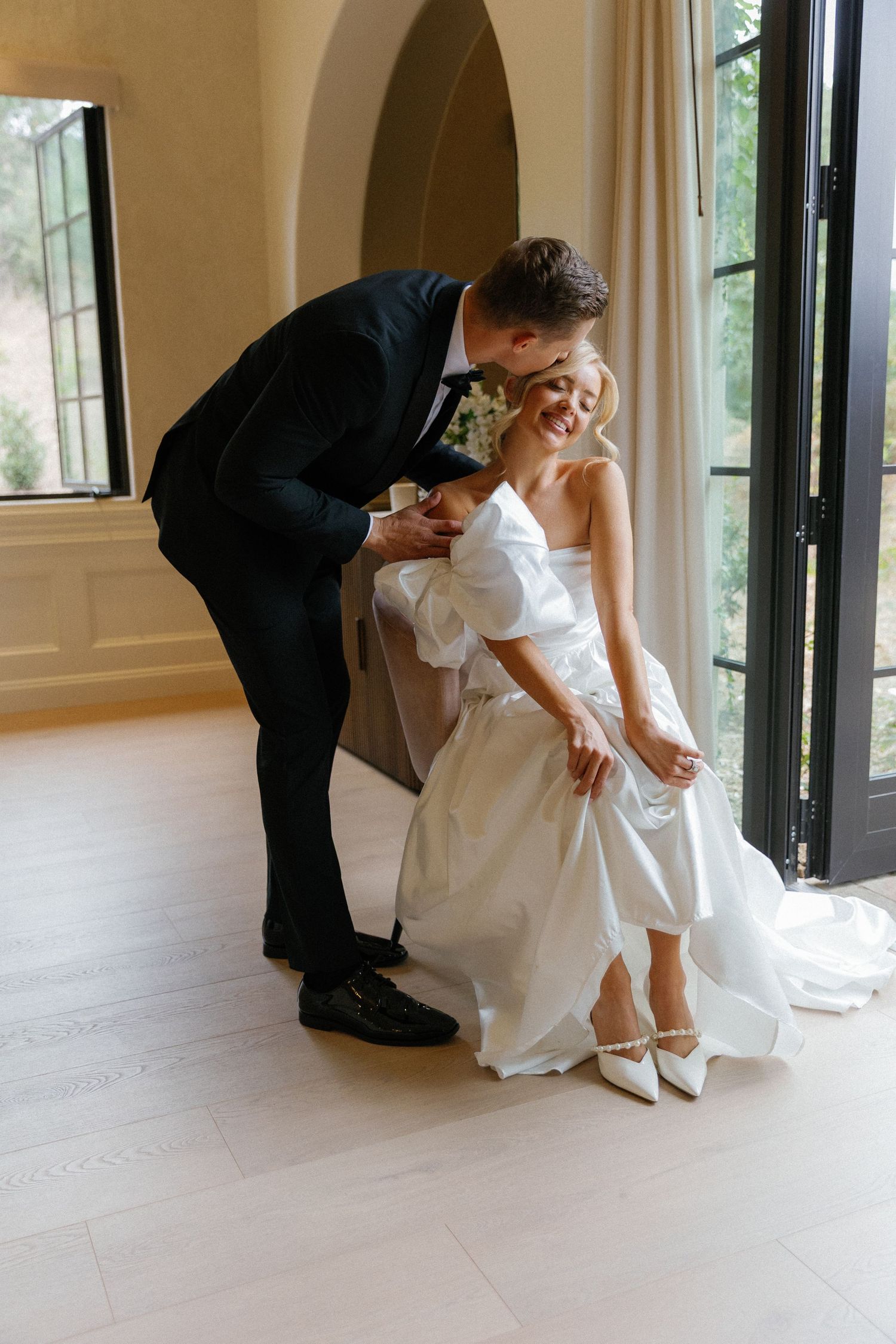 Father in black suit helps daughter in white wedding dress put on her shoes in elegant room with arched doorway.