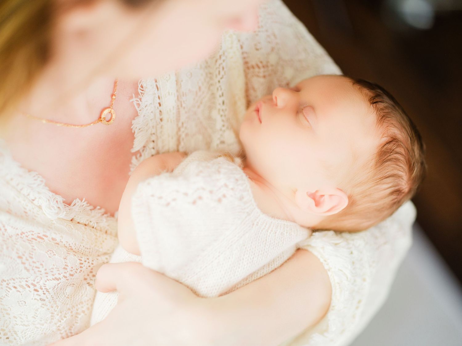 A sleeping newborn baby dressed in white lace clothing lies peacefully on a soft white blanket.
