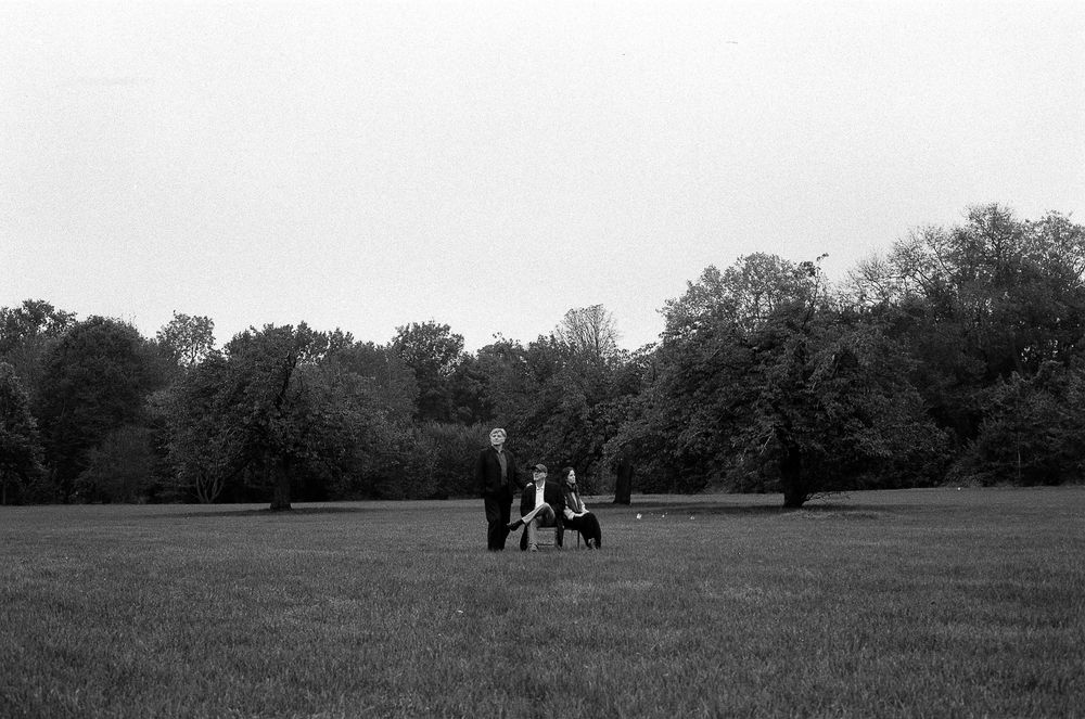 A serene black and white scene shows distant figures walking with a dog across a vast grassy field bordered by trees.