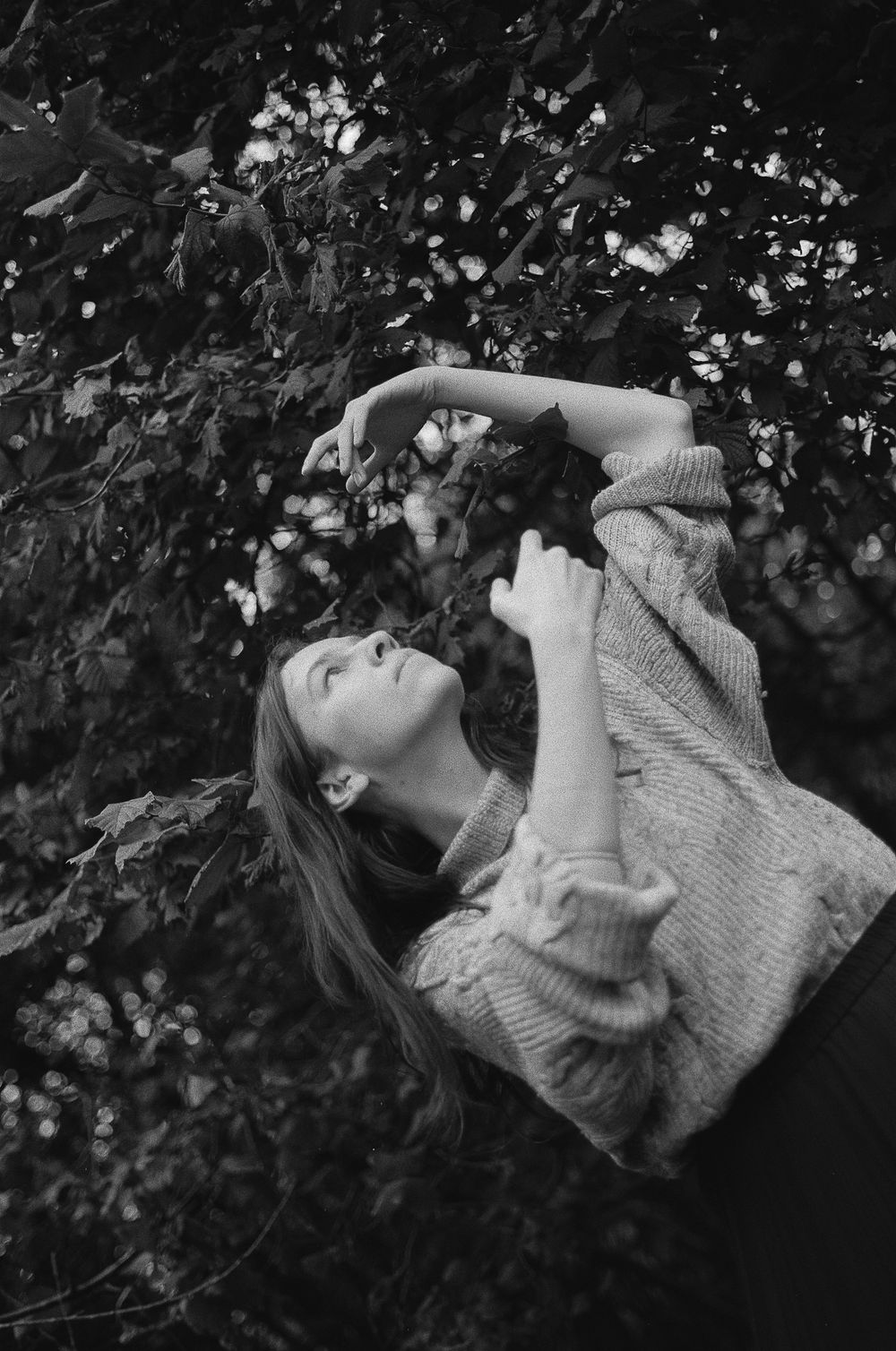 A person in a sweater reaches upward among tree branches in a dreamy black and white photograph.