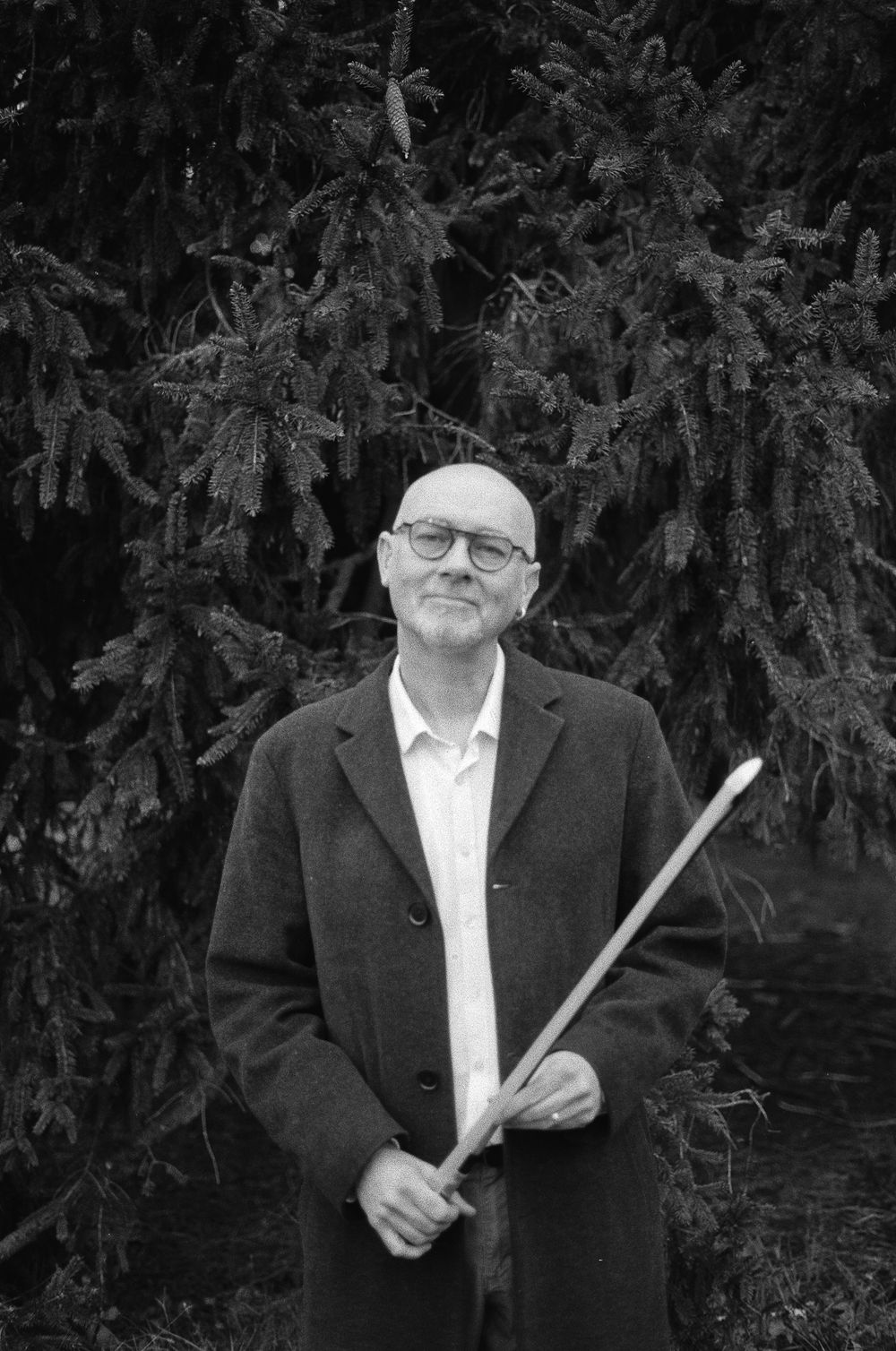Black and white portrait of a person in glasses and suit jacket holding a conductor's baton near evergreen trees.