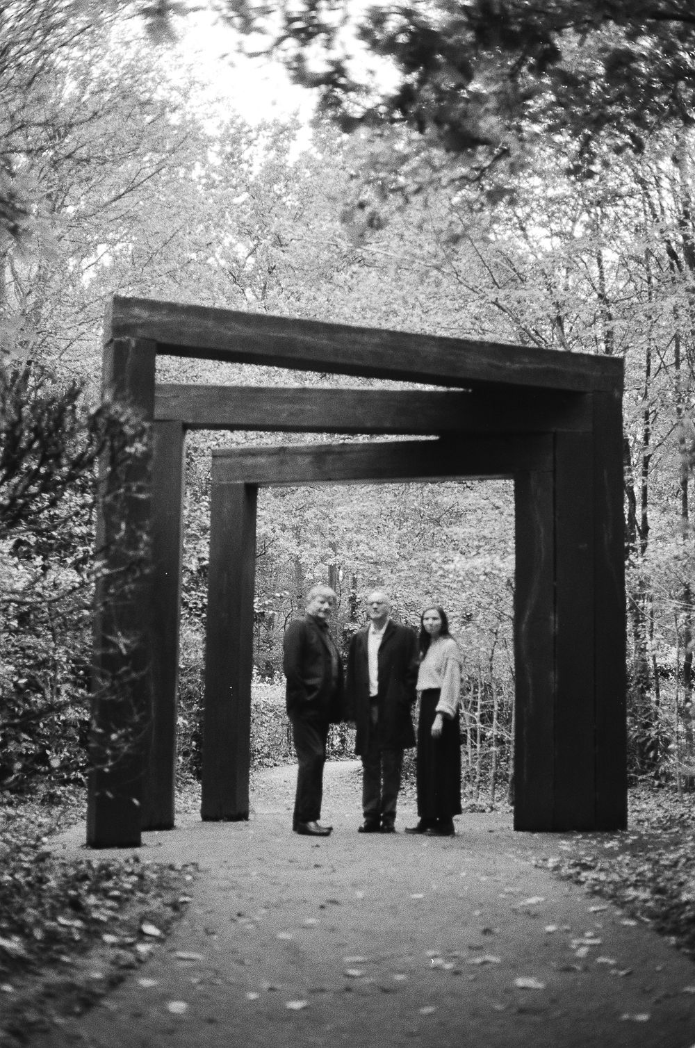 Black and white photo of people standing beneath a wooden gateway structure along a forest path.