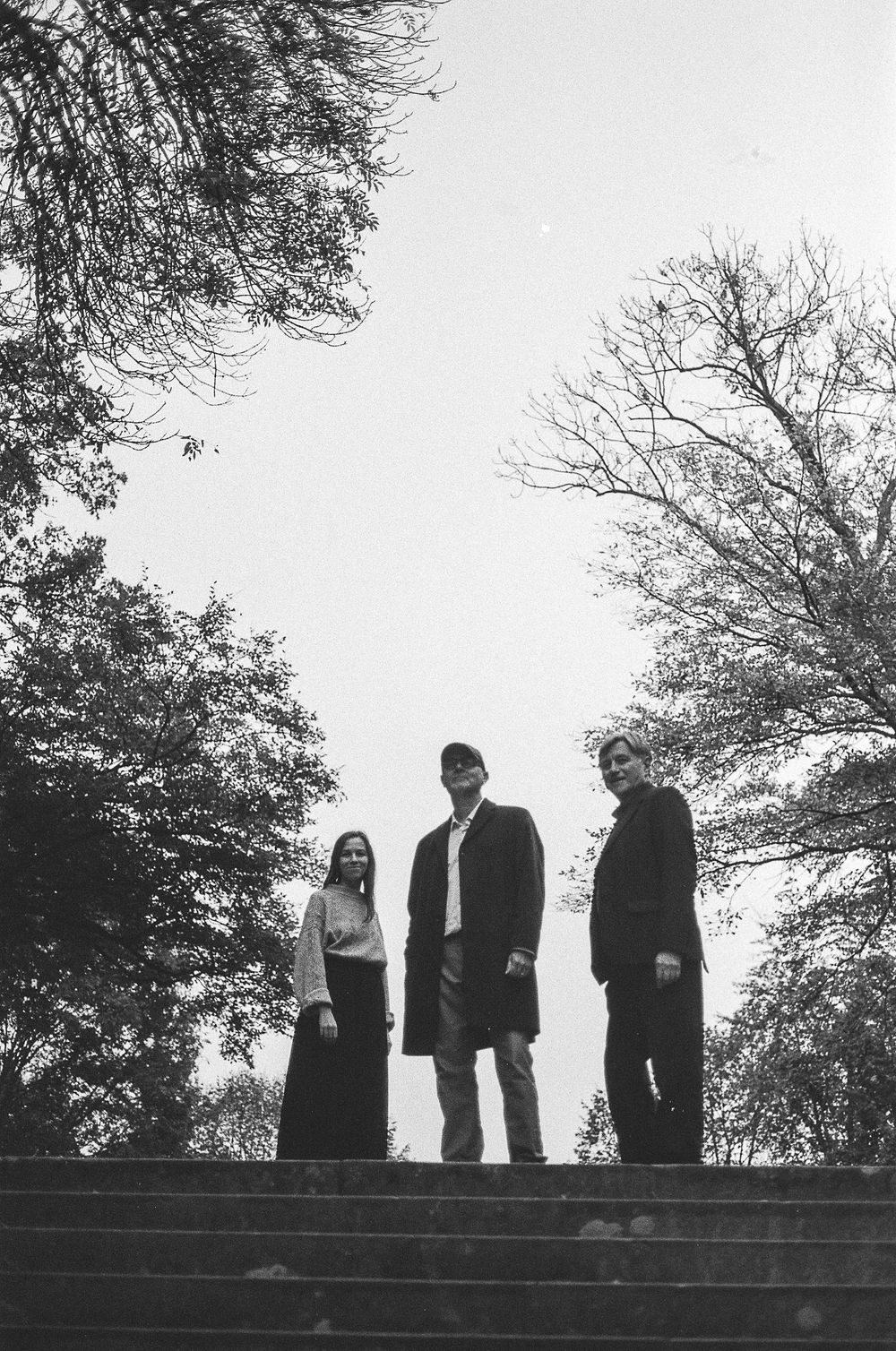 Dramatic black and white photograph of three persons standing at the top of stone steps surrounded by bare trees.