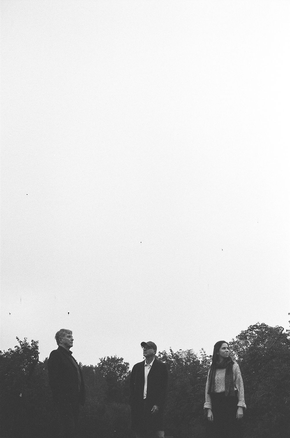 Three people stand outdoors in a black and white photograph taken in a park or wooded setting in low light.