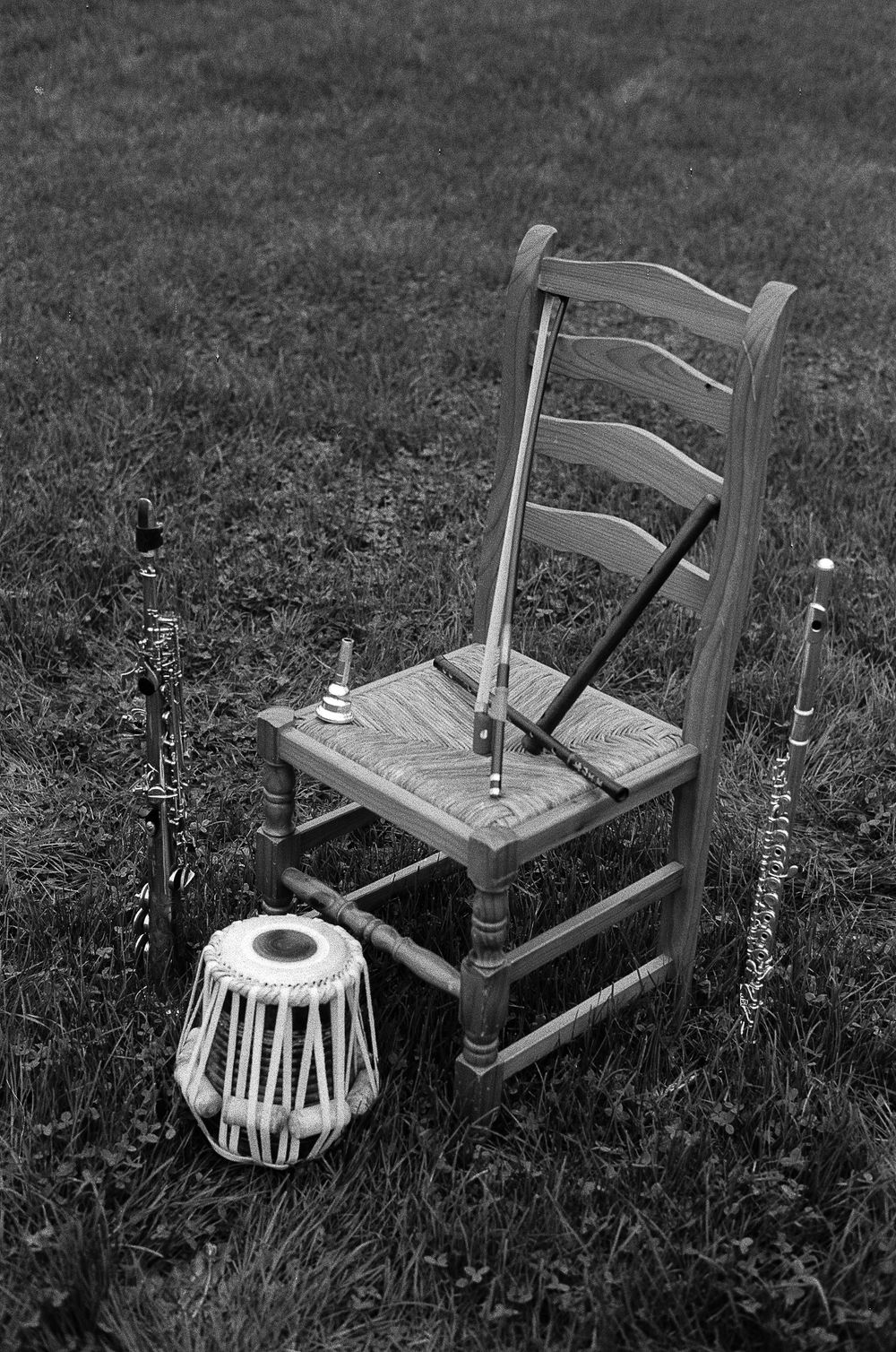 Black and white photograph showing a wooden ladder-back chair and tabla drum on grass with musical instruments around them.