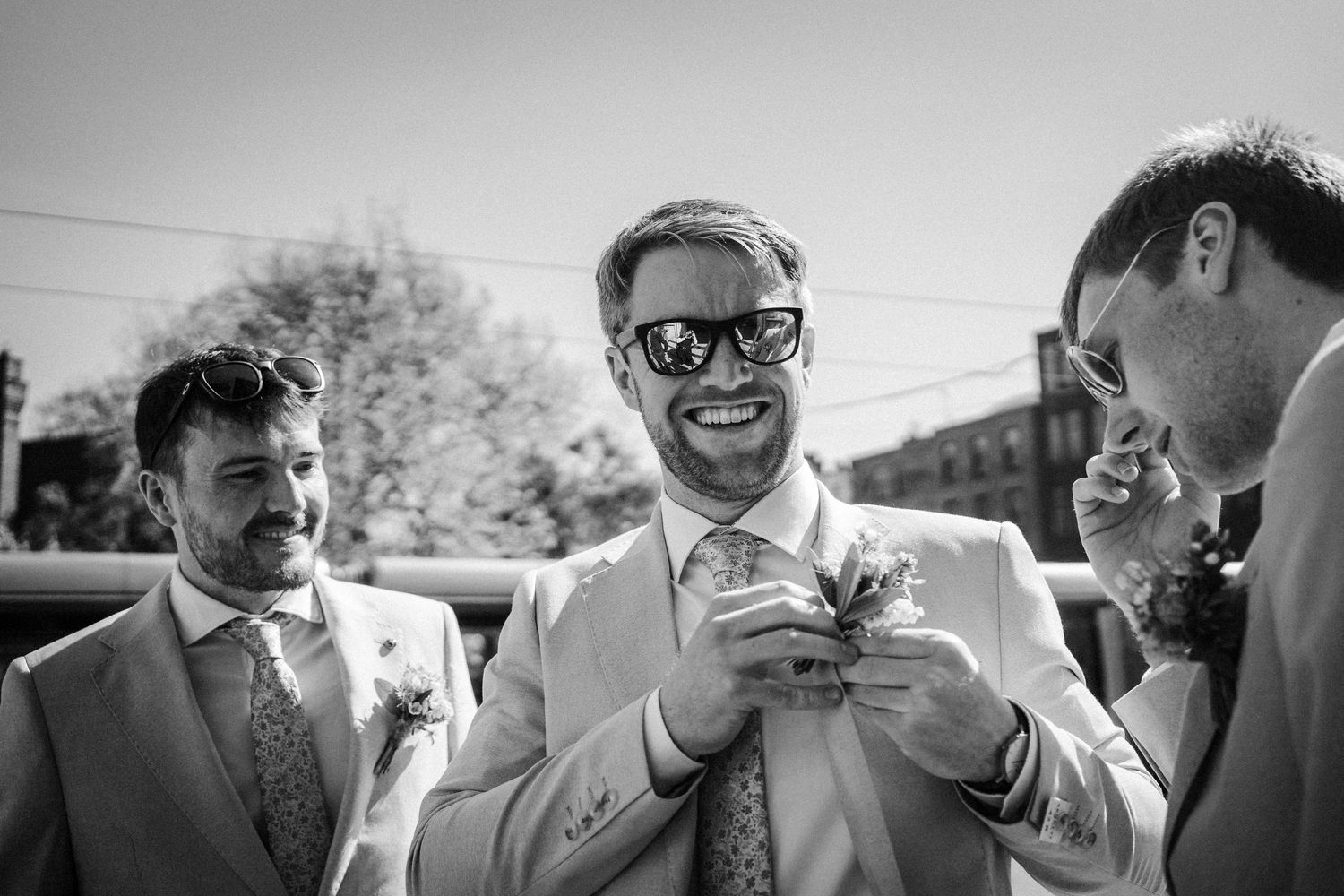 A candid black and white photo of three men in suits sharing a lighthearted moment at an outdoor event.