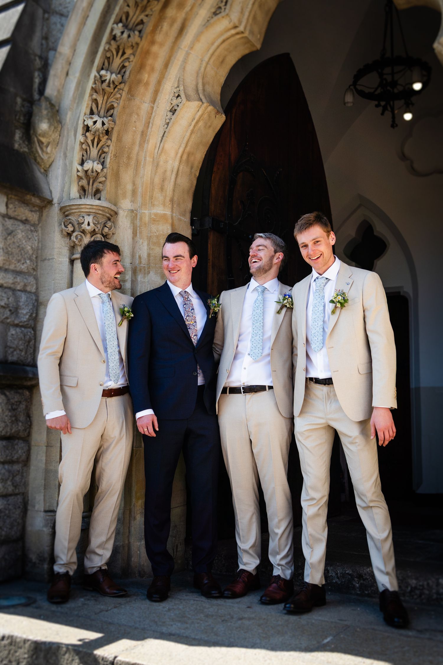Four groomsmen in light suits and ties stand together smiling under a stone archway at an elegant venue.