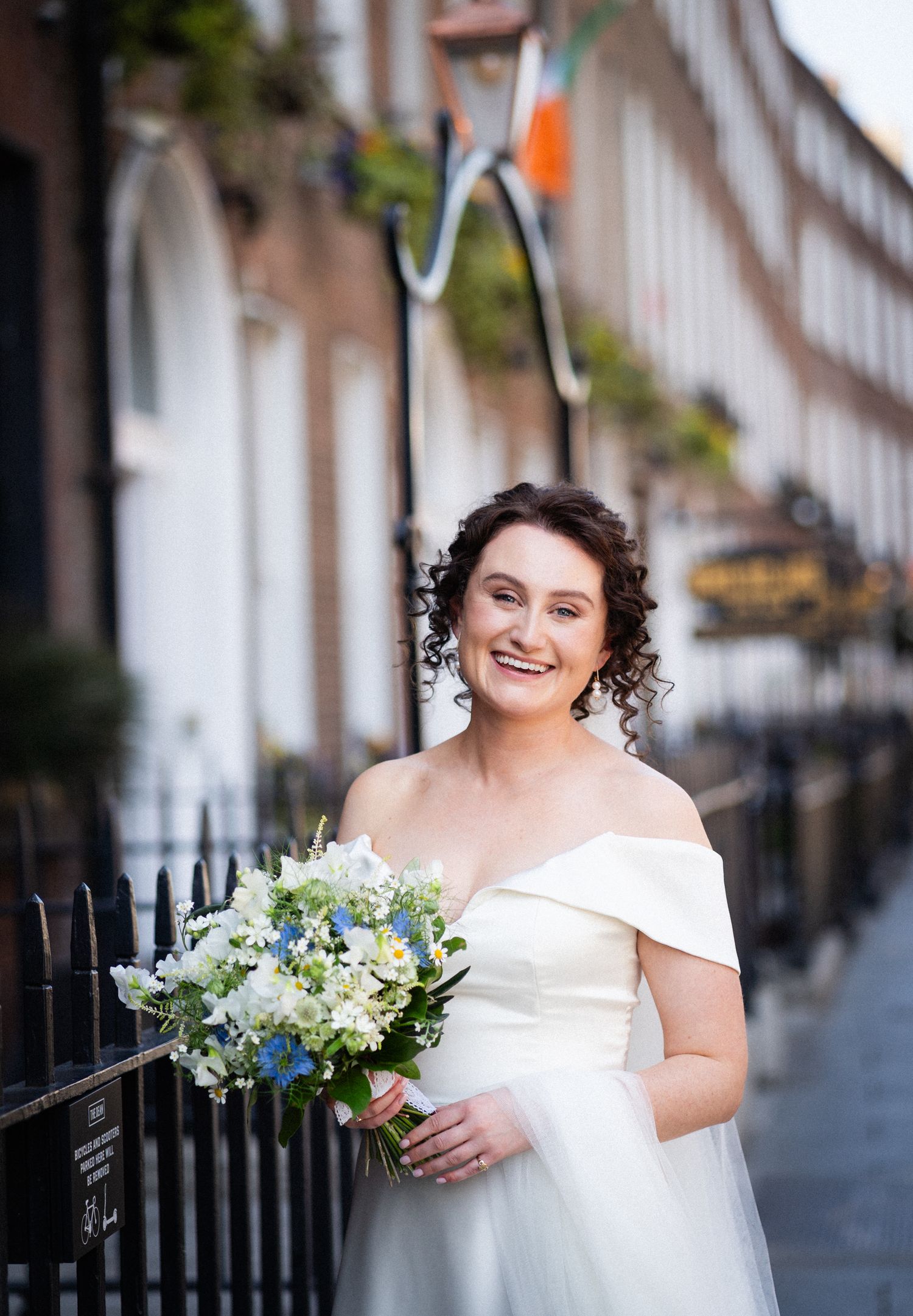 A bride in an off-shoulder white wedding dress holds a beautiful bouquet of blue and white flowers on a Dublin street.