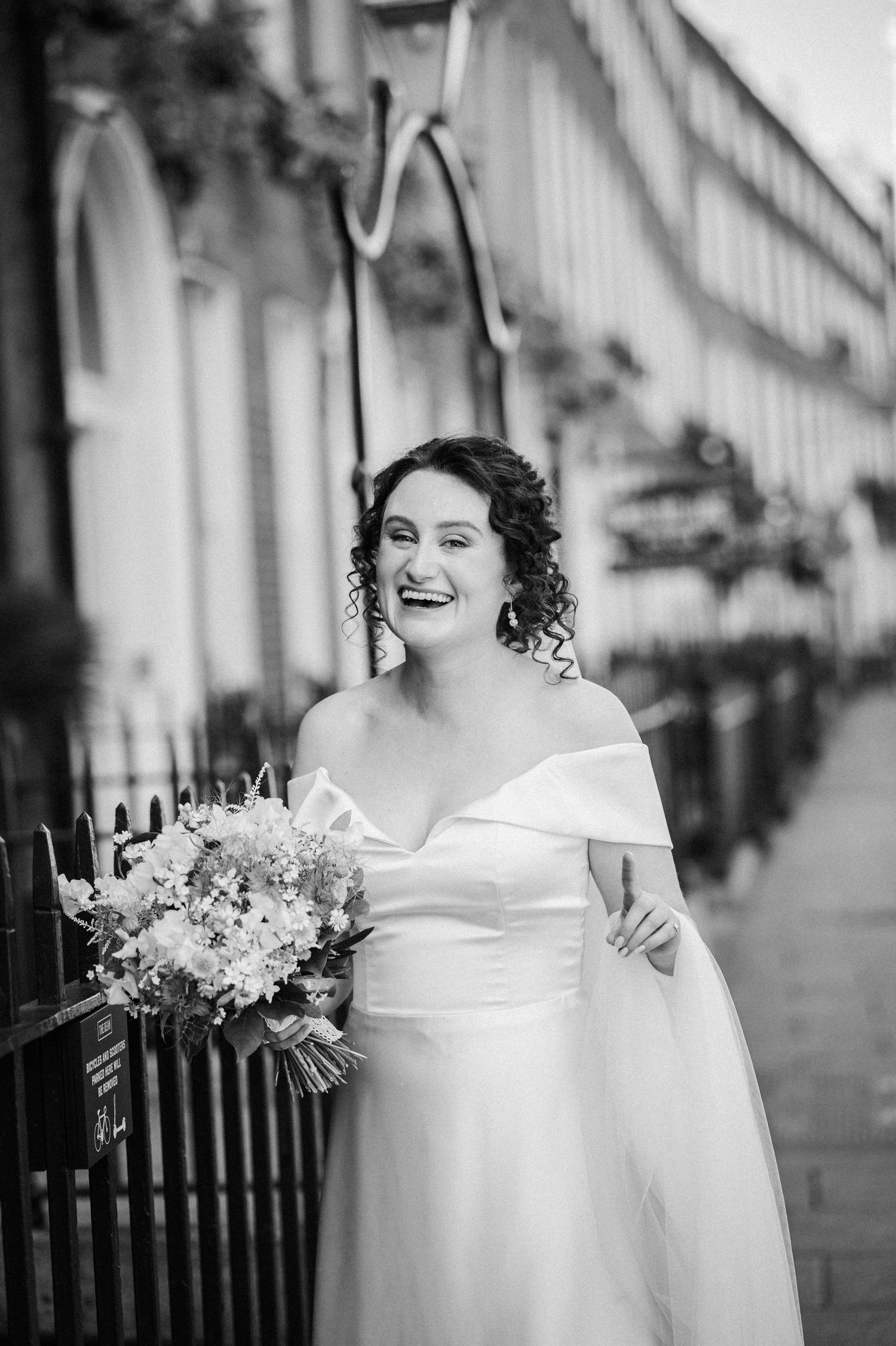 A bride wearing an off-shoulder white wedding gown holds a bouquet while standing by a wrought iron fence.