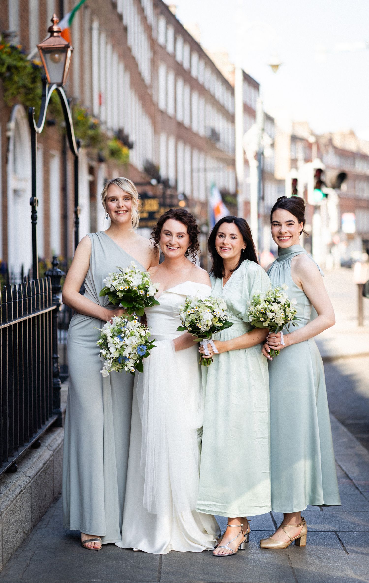 Wedding party in pale mint and silver dresses pose together holding delicate white floral bouquets on a city street.