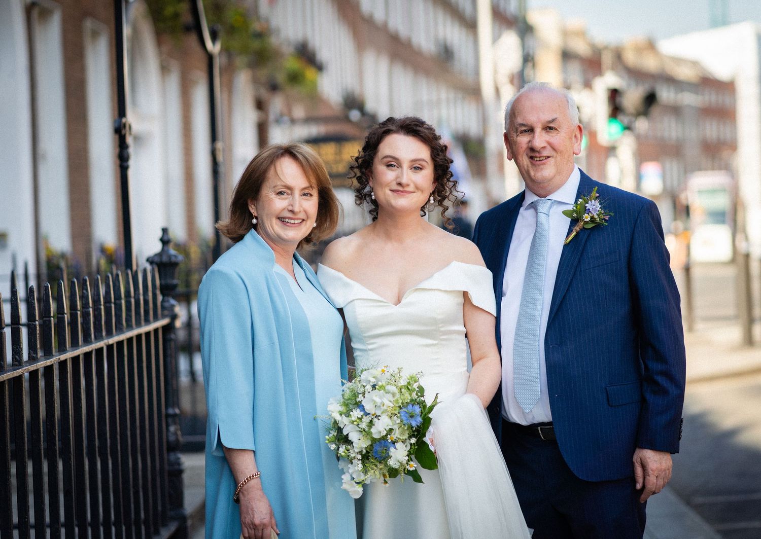 Three people in formal attire pose together outdoors on a sunny day with one holding a white and blue bouquet.