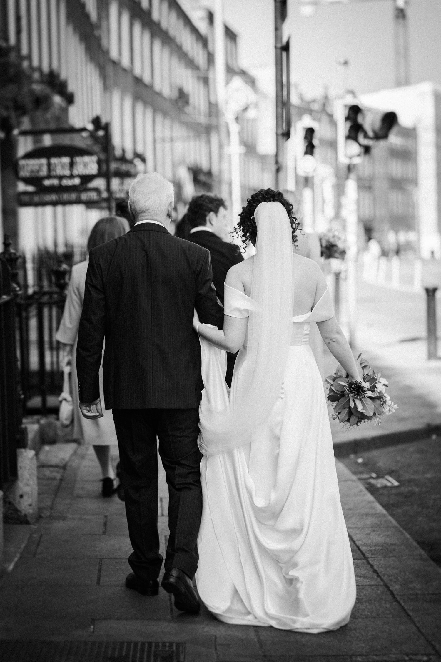 Black and white photo of a bride in a flowing white dress walking down a city street with two companions.