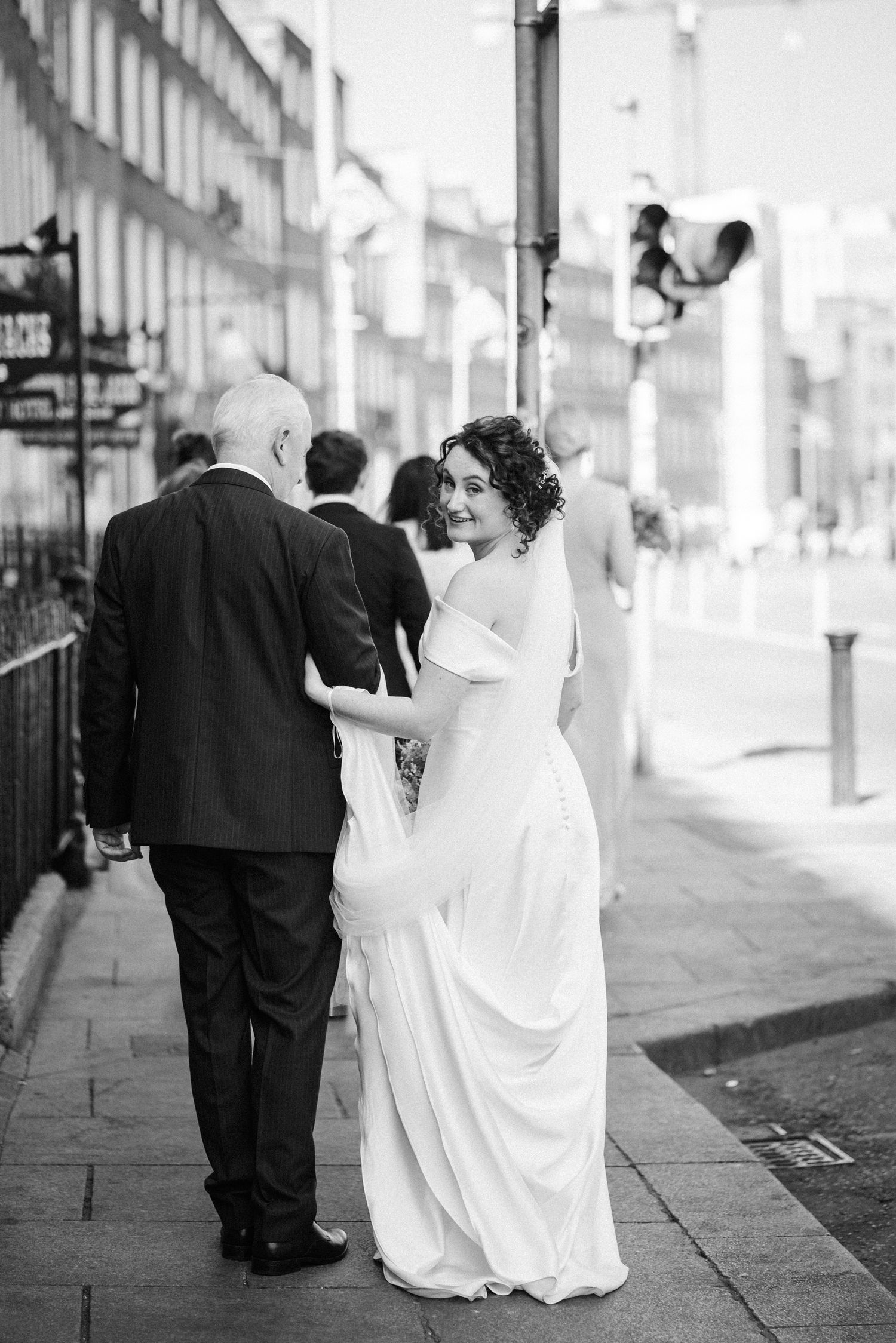 A black and white photograph of a bride in a flowing white dress walking down a city street with companions.